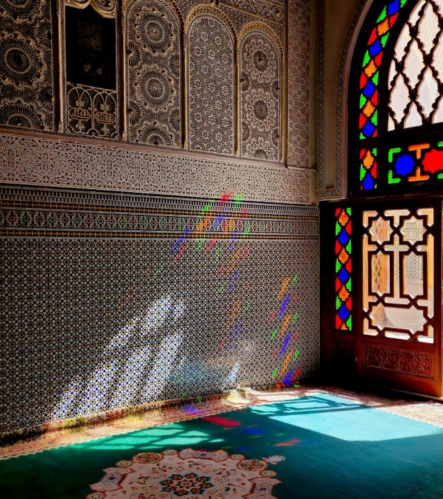 Colorful stained glass window casting rainbow light on ornate Moroccan mosque interior.
