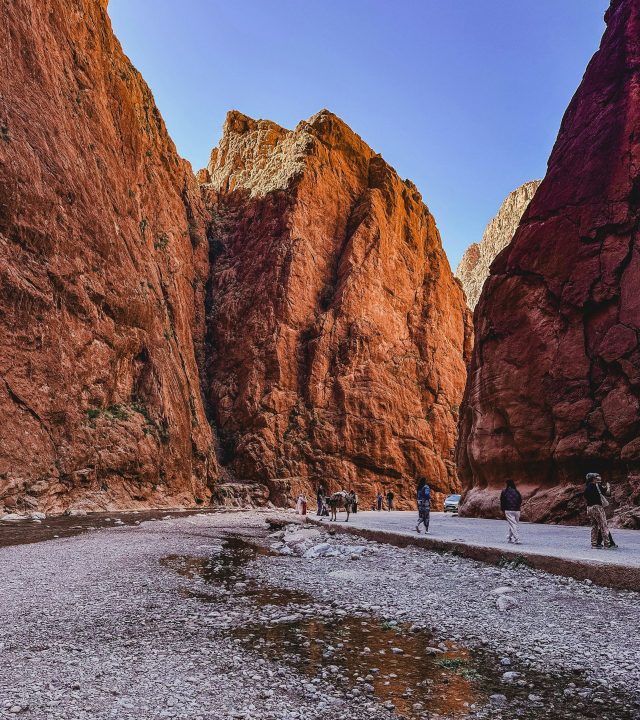 Sunlit red rock walls of Todra Gorge with people exploring the scenic path.
