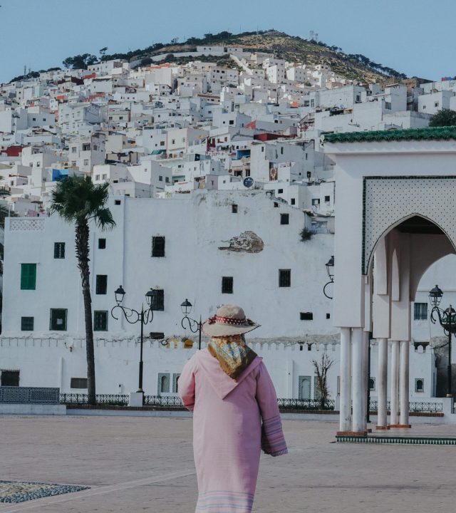 A woman in traditional attire in Tetouan, Morocco, with hillside architecture in the background.